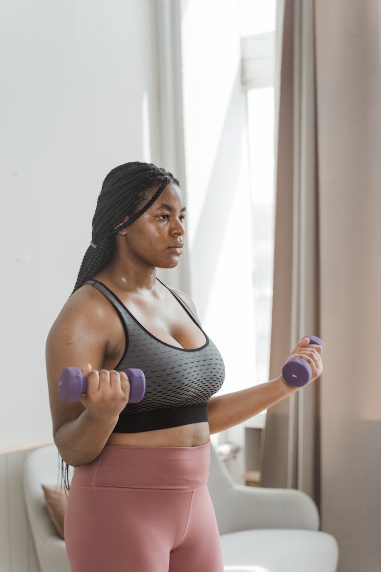 Curvy woman with braided hair working out with dumbbells indoors, embracing body positivity.