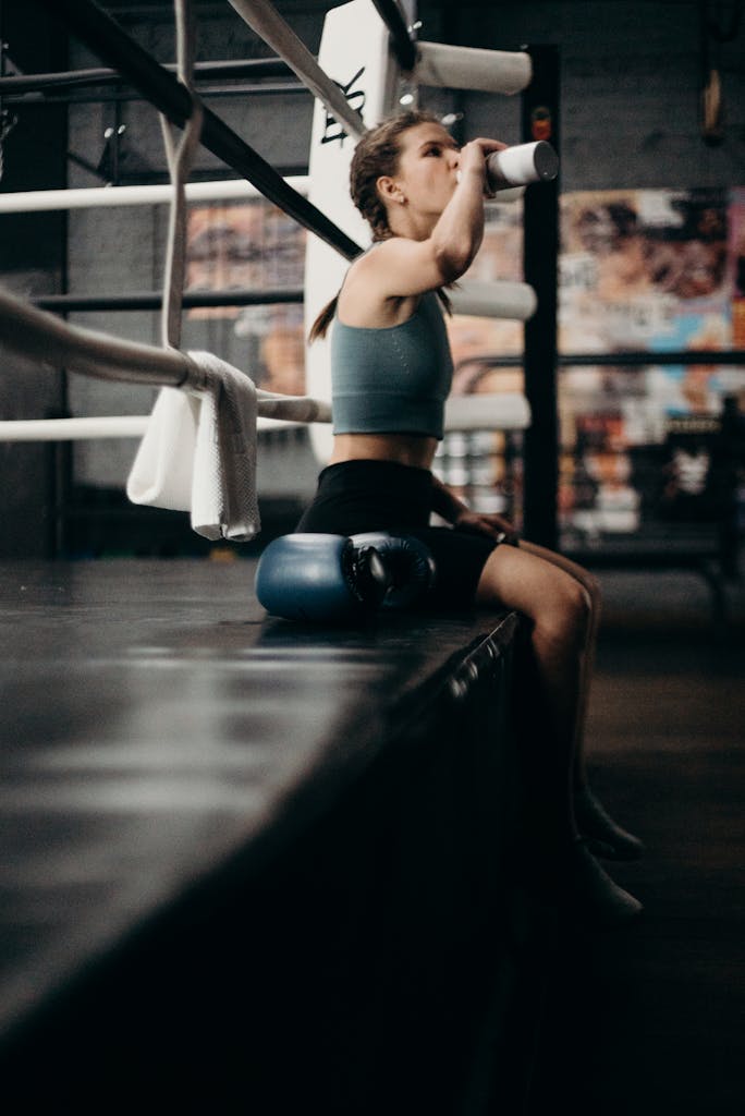 Woman boxer drinking water in gym, sitting on edge of boxing ring.